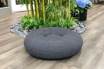 Dark gray circular floor cushion sits on light brown wooden floor.  Plants, bamboo, and stones surround it