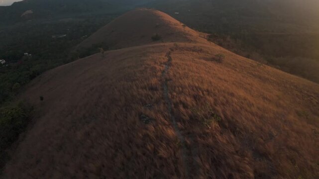 Forward drone sweep traces a single footpath over sun-bathed golden grass hills of Mount Tapyas, revealing tranquil forested lowlands around Coron Island bathed in warm Philippine dawn light.