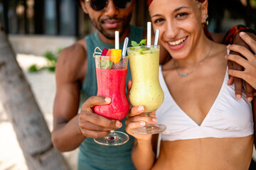 Smiling couple holding colorful tropical smoothies on a sunny beach, enjoying refreshing drinks and vacation time together in a relaxed, tropical setting