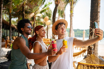 Group of cheerful young friends taking a selfie while holding colorful tropical drinks at a beach resort, capturing joyful summer memories