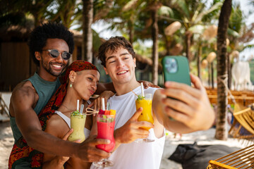Group of cheerful young friends taking a selfie while holding colorful tropical drinks at a beach resort, capturing joyful summer memories