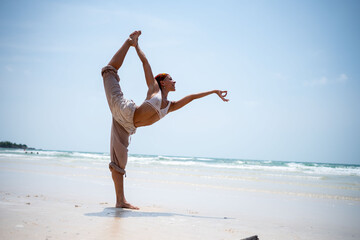 Woman with a headscarf performs a standing yoga pose on a tropical beach, balancing gracefully with...