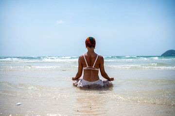 Woman practicing yoga on a tropical beach, sitting in lotus pose with her back to the ocean,...