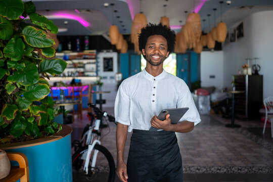 Young restaurant owner or manager holding a digital tablet in a modern, stylish bar or cafe, standing confidently and smiling in a welcoming hospitality setting