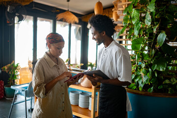 Smiling waiter and guest completing a mobile payment transaction inside a stylish, tropical-themed...