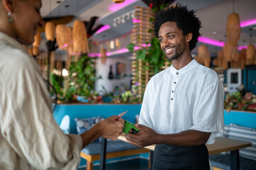 Smiling waiter and guest completing a mobile payment transaction inside a stylish, tropical-themed...