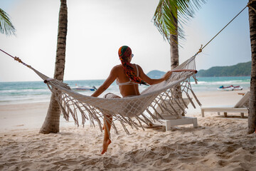 Woman sitting on a hammock between palm trees on a sandy beach, gazing at the ocean, enjoying...