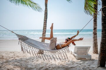Playful woman lying in a hammock on a tropical beach, stretching her arms and legs joyfully under palm trees with the ocean in the background.