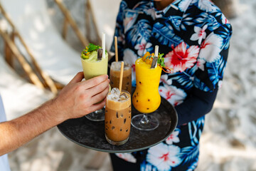 Tropical drinks being served on a tray at a beachside cafe, with a customer reaching for a...