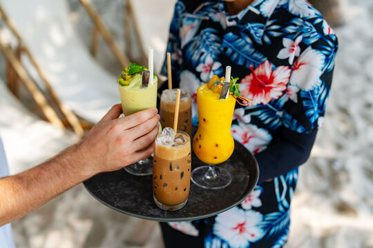 Tropical drinks being served on a tray at a beachside cafe, with a customer reaching for a smoothie. Vibrant and refreshing beverages in a relaxed, sunny setting