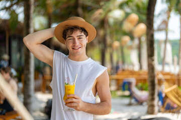 Smiling young man in a hat holds out a tropical mango drink in focus, offering it cheerfully on a...