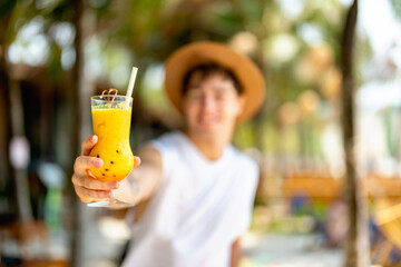 Smiling young man in a hat holds out a tropical mango drink in focus, offering it cheerfully on a...