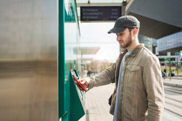 Young man validating tram ticket using mobile phone app at automatic ticket machine in train station