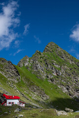 Mountain hut at the base of rocky peaks in the Fagaras Mountains, Romania. Green alpine slopes in the Carpathians under clear blue sky.