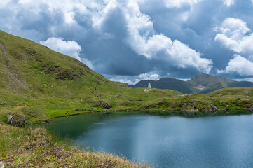 Capra Lake in the Fagaras Mountains, Romania. Cloudy sky over a calm alpine lake and green mountain slopes in summer.