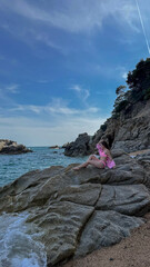 A slim woman in a swimsuit sitting on coastal rocks at Playa Cala Sa Boadella, Lloret de Mar, Spain. Relaxed summer vibe with copy space for travel ads.