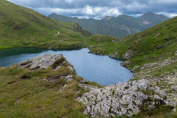 Mountain lake surrounded by green hills and rocky slopes in the Fagaras Mountains, Romania. Cloudy summer weather in the Carpathians.