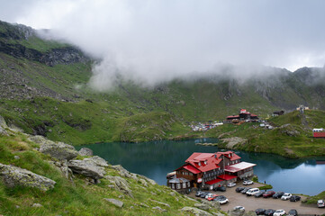 Bâlea Lake in the Făgăraș Mountains, Romania, with mountain lodges, parked cars, tourists, and cloudy weather. High-angle view of the glacial lake surrounded by green alpine slopes and dramatic peaks.