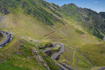 Curvy section of the Transfagarasan Road in the Carpathian Mountains, Romania. Aerial view of winding mountain road with cars and dramatic green slopes on a clear summer day.