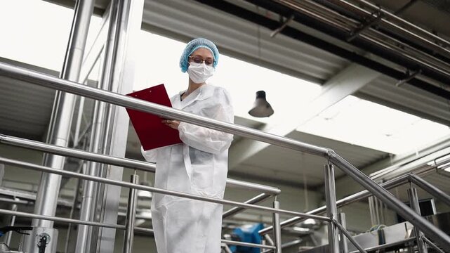 Female worker in protective workwear working in medical supplies research and production factory. She is standing or walking on control bridge and watching at production line.