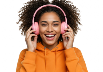 An excited young woman with curly hair, wearing an orange hoodie, smiles broadly while listening to music with pink headphones, isolated on a transparent background