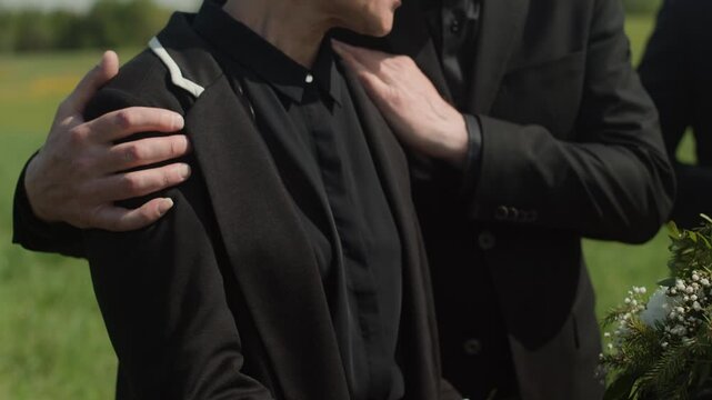 Tilt up cropped slowmo of unrecognizable widow in mourning touching wooden coffin sorrowing over death of loved one and being gently supported by close friend or relative at outdoor funeral