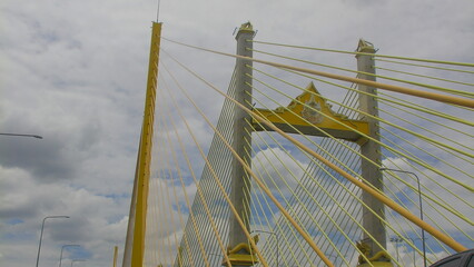 Towering structure of Bhumibol Bridge captured from below, emphasizing the tall suspension pillars and radiating yellow cables beneath an overcast sky.