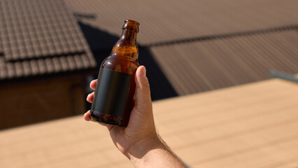 Hand holding a blank beer bottle against a rooftop background for advertising mockup. Concept of...