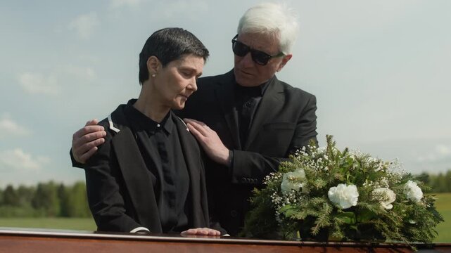 Medium shot of mature Caucasian widow in mourning looking at coffin at outdoor funeral ceremony and being supported by senior man in black suit sorrowing together over death of loved one