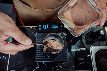 Spoonful of chocolate or cocoa powder being added into a milk-filled metal pitcher during the...