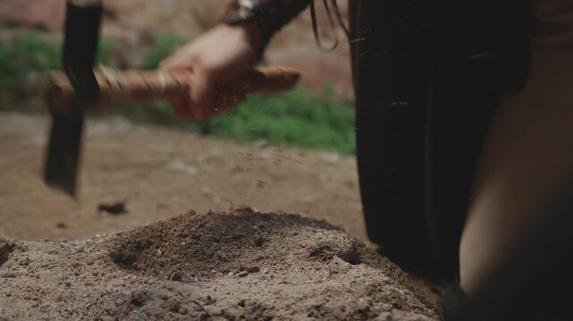 Close-up of a hand in a leather bracer digging soil with an ancient pickaxe. Historical reenactment or fantasy scene of hard labor, excavation, or searching for treasure.