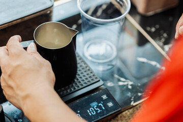 Close-up of milk being weighed in a metal pitcher on a digital scale at a coffee stand before...
