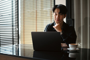 A Professional Asian man works intently on a laptop with a cup of coffee beside him in a sunlit room