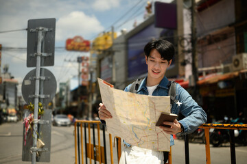 A Young Asian man holds a paper map and smartphone, standing on a lively street, navigating his way...