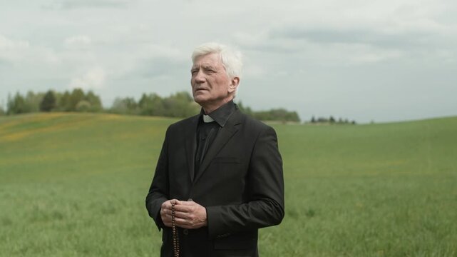 Medium shot of serious mature Caucasian priest wearing black soutane and holding rosary beads looking up and praying at outdoor funeral or memorial service
