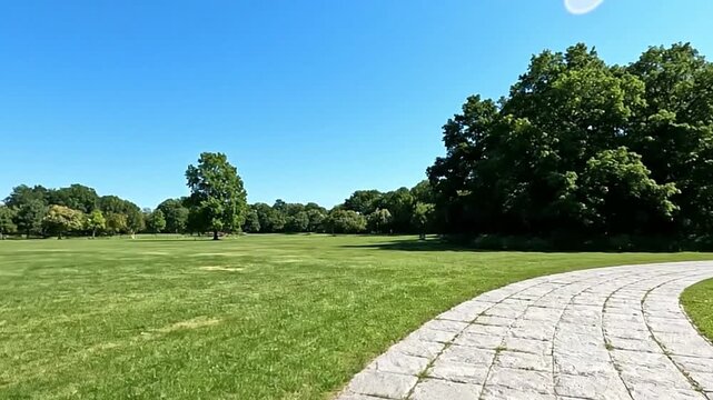 A wideopen grassy park under a blue sky bordered by trees and a curved stone path