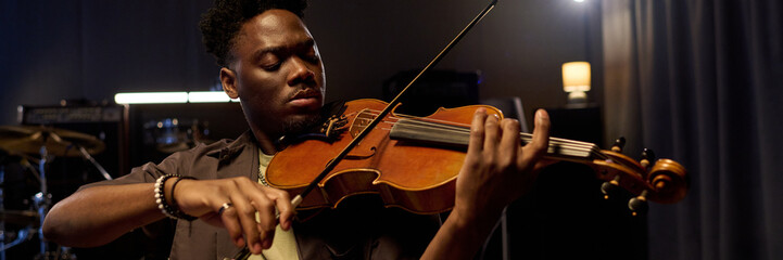 Black young adult man playing violin with focused expression on stage, holding instrument under chin and drawing bow across strings, performing music in professional studio setting © pressmaster