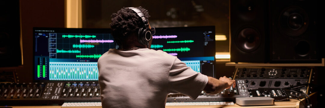 Black man young adult sitting at mixing console operating audio equipment in professional recording studio, wearing headphones and working on music production