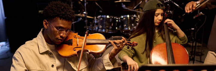 Young Black man playing violin and young Latin woman playing cello in music studio, both focused on...