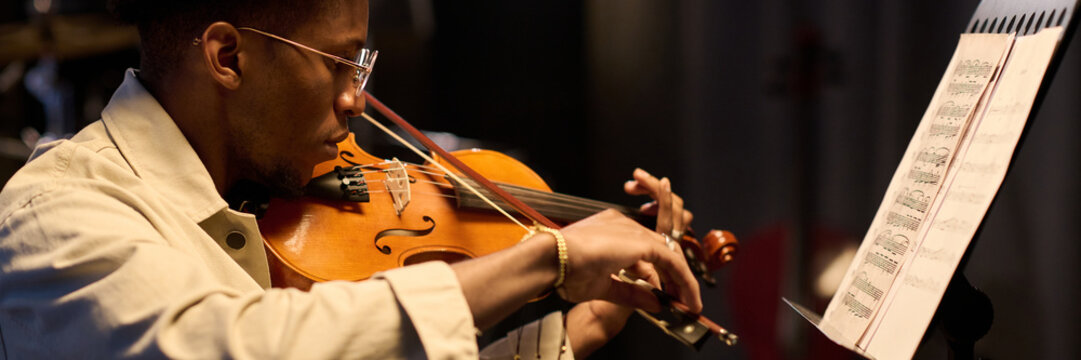 Young adult Black man playing violin while reading sheet music on stand, sitting indoors with focused expression, wearing glasses, demonstrating musical performance skills