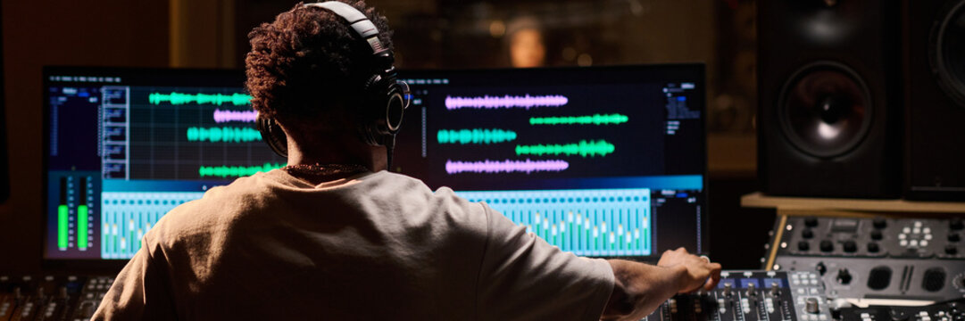 Black man wearing headphones working at audio mixing console in professional recording studio, adjusting sound levels on large digital display with colorful waveforms visible