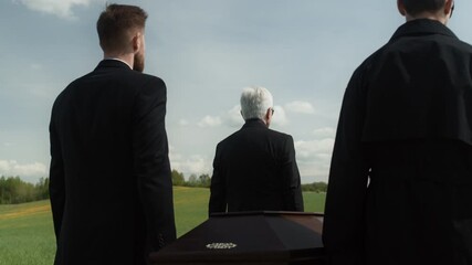Rear view shot of unrecognizable men wearing black suits carrying wooden coffin along cemetery helping as pallbearers during funeral ceremony