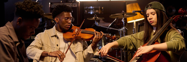 Black young adult man playing violin next to Latin young adult woman playing cello and Black young...