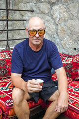 Middle-Aged Man in Yellow Glasses Holding Turkish Coffee on Red Oriental Sofa