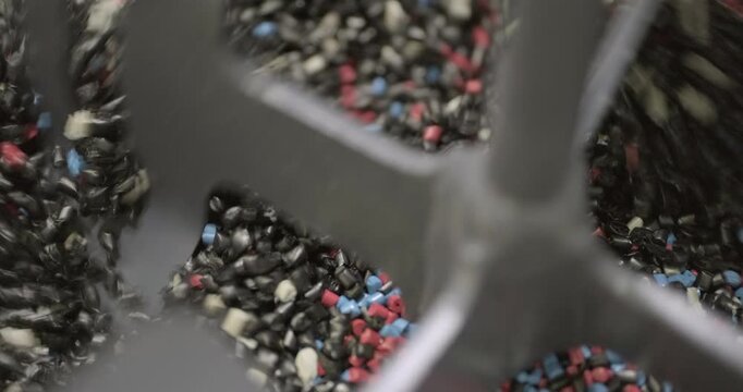 Pieces of multi-colored plastic are mixed in an industrial mixer before being disposed or reused. Close up handheld shot, real time speed, artificial indoors lighting.
