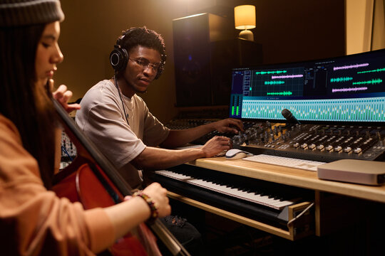 Young Black man wearing headphones operating audio mixing console and computer while young Latin woman playing cello in music recording studio, both focused on production process