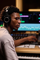 Young adult Black man wearing headphones working at audio mixing console in music studio, operating...