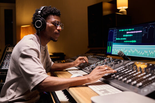 Young adult Black man wearing headphones operating audio mixing console and computer monitors in professional recording studio, adjusting sound levels and working with music production equipment - Powered by Adobe