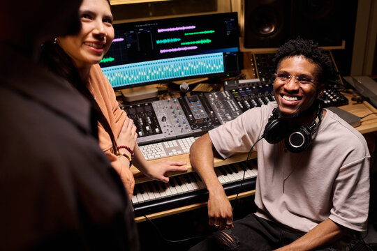 Young Black man sitting at music production workstation smiling at multiethnic group, wearing headphones around neck, engaging with colleagues in professional recording studio environment