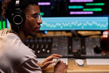 Young adult Black man wearing headphones operating audio mixing console in professional recording...